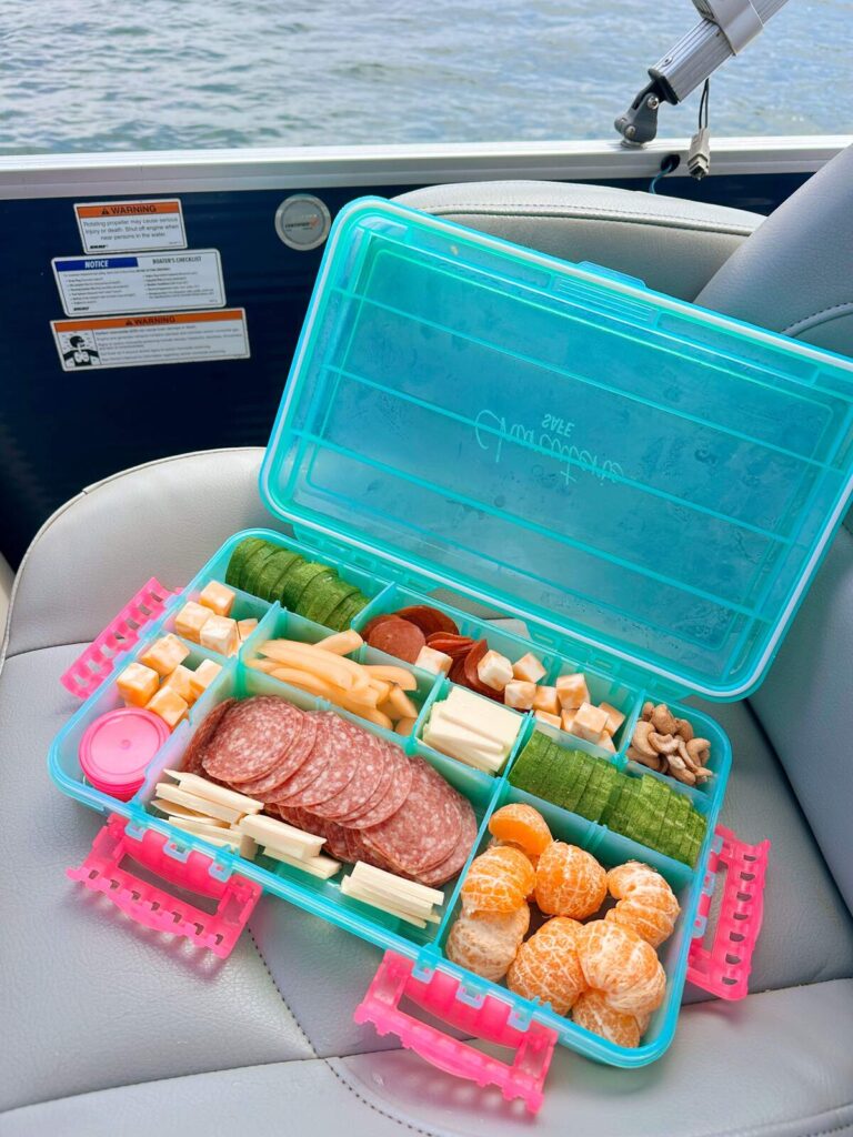 A snackle box sitting on a seat in a boat filled with meats, cheeses, fruits and nuts. This is one of our favorite ways to eat lunch on a boat.