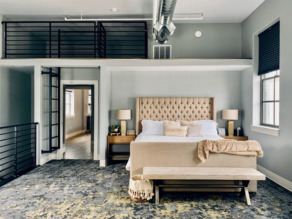 Airbnb bedroom with stunning architectural features such as iron railing, a cutout in the wall for a bd, and exposed pipes.