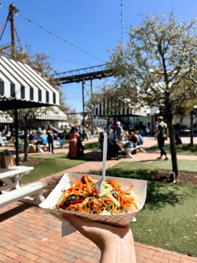 This is me holding up my delicious chicken bulgogi meal from Club Sandwich- my favorite food truck at the Silos. The background shows the shaded picnic tables where you can sit and eat.