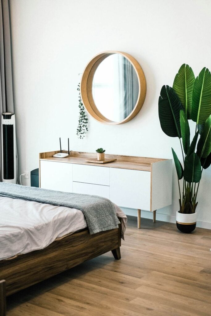 A corner of a bedroom featuring a white dresser, round wood mirror, and large plant.