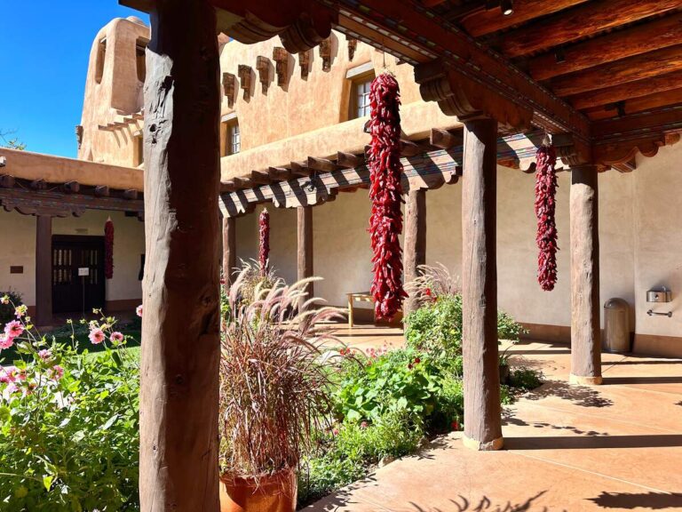 The courtyard of the New Mexico Museum of Art. It is one of my favorite spots in Santa Fe! It features hanging ristras, art, flowers, and adobe architecture.